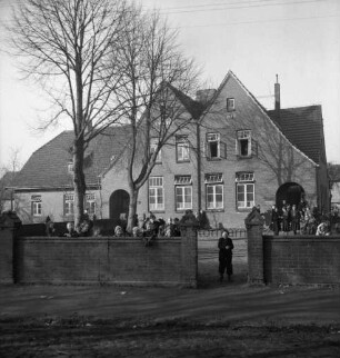 Volksschule: Hauptstraße: vorn Mauer: dahinter Schulkinder auf Schulhof: rechts im Durchgang Junge: links Bäume: hinten Schulgebäude
