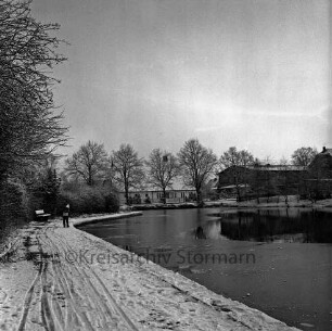 Dorfteich: links Schulkind am Rand: hinten Bäume und Häuser: Winter