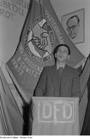 Ein Mann am Rednerpult in der Landesproduktionsgenossenschaft "Otto Buchwitz", im Hintergrund ein Portrait Otto Grotewohls