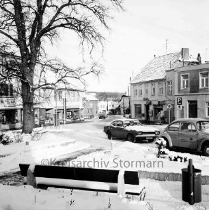 Paul-von-Schoeneich-Straße: Blick von Rathaus Richtung Ahrensböker Straße: links Einmündung von Bergstraße: im Winter: im Vordergrund Grünfläche mit Sitzbank, Baum, Papierkorb: auf Straße Autos