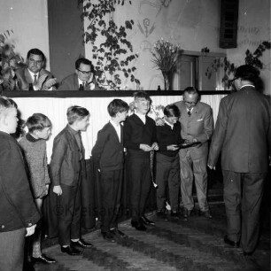 Kreisfußballverband Stormarn: Verbandstag: Reinfeld: Festsaal des "Hotel Stadt Hamburg": Ehrung der Meister: Vorsitzender Erwin Gesche bei Ehrung einer Gruppe von Jungen: dahinter Verbandsleitung auf Podium, klatscht: Grün- und Blumenschmuck, rechts hinten Tür, 22. Mai 1965