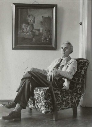 Porträt Oskar Schmidt, Ansicht in einem Sessel unter einem Gemälde (Zwei Mädchen mit Teddy vor einem Kasperletheater)