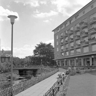 Grünanlage mit Fußweg, Jägerzaun und Straßenlampe: links Trave: rechts Wohnblock: hinten links Brücke an Hagenstraße: dahinter links Gebäude