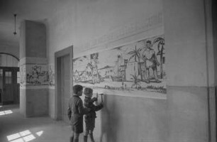 Dekorative Ausgestaltung der Klassenräume und Schulflure in der Einheitsschule Baumschulenweg und Oberschule Niederschöneweide in Vorbereitung des Deutschlandtreffens der Jugend für Frieden und Völkerfreundschaft zu Pfingsten 1950 : Schüler beim Anbringen eines Plakats zum Deutschlandtreffen an einer Wand im Hausflur einer Schule