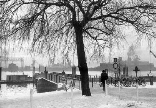 Hamburg-Flottbek. Der Fähranleger in Teufelsbrück, dem Mündungsbereich des Baches Flottbek in die Elbe, im Winter. Im Hintergrund das Werftgelände der Deutschen Werft. Passagiere laufen zur Fähre