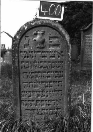 Grabstein 400 (Index: 100102823) Material: Sandstein Zustand: leicht verwittert Maße HxBxT: 135x57x8 cm Beschreibung: Stele, Rundbogenabschluss, im Bogen plastische Levitenkanne, Schriftfeld eingetieft Symbolik: Levitenkanne: Grabstein 400