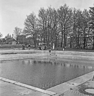 Freibad: vorn Schwimmbecken: dahinter Arbeiter mit Schaufel und Trecker bei Außenarbeiten: hinten links Sportplatz: hinten rechts Bäume und Häuser