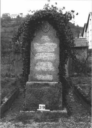 Grabstein 39 (Index: 99805053) Material: ??? Maße HxBxT: 190x65x28 cm Beschreibung: Stele auf gestuftem Sockel mit Rundbogen, efeubewachsen Symbolik: Blüte im Rundbogen: Grabstein 39