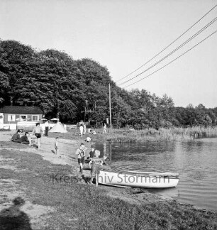 Lütjensee: Campingplatz am Nordstrand: rechts See: vorn Jungen mit Ruderboot: links dahinter Personen: hinten links Holzhaus mit Gastronomie und Toiletten