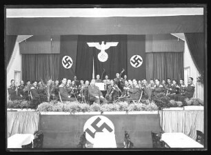 Musikkapelle in Reichsuniform auf Bühne sitzend. Rückwand Reichsadler