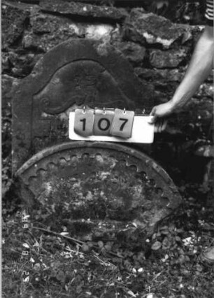 Grabstein 107 (Index: 100110753) Material: Sandstein Zustand: Giebelfragment, stark verwittert Maße HxBxT: 54x82x20 cm Beschreibung: Rundbogenabschluß (Fragment): Grabstein 107