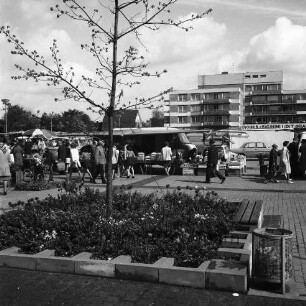 Wochenmarkt: Rathausplatz: Marktwagen, Marktstände, Kunden, Autos: im Vordergrund Blumenrabatte mit Bäumchen, Parkbank, Papierkorb: hinten rechts Wohn- und Geschäftsblock der nördlichen Randbebauung, mit Geschäft "Johs. Schmidt Markt"