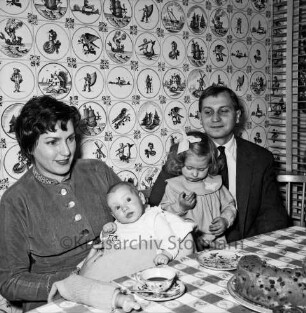 Juscha und Siegfried Schneider-Döring: Kunstkeramiker: mit Baby und Kleinkind: am gedeckten Tisch, Kuchen: dahinter Kachelwand mit holländischen Motiven