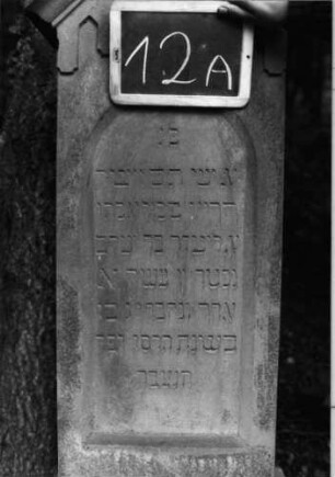 Grabstein 12 (Index: 100090870) Material: Sandstein Zustand: leicht verwittert Maße HxBxT: 157x42x18 cm Beschreibung: Stele, getreppt. Sockel, giebelart. Abschluß mit Verdach. , Schriftfeld m. Rundbogenabs Symbolik: gestelztes giebelartiges Fries: Grabstein 12A