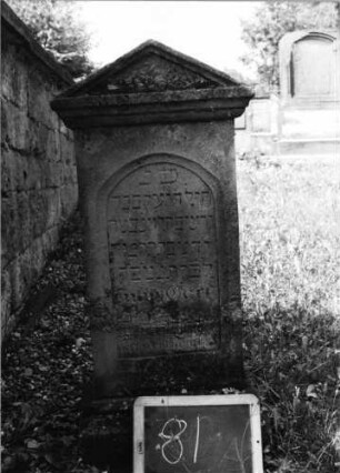 Grabstein 81 (Index: 99801929) Material: Sandstein Zustand: stark verwittert mit Textverlust Maße HxBxT: 86x36x17 cm Beschreibung: Stele, profilierter Sockel, Giebel, eingetieftes Schriftfeld mit Rundbogen Besonderheit: Kindergrab: Grabstein 81