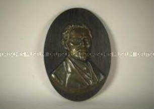 Brustbild von August Bebel, Messingrelief mit Bebelzitat auf einem ovalen Holzbrett, dunkelbraun, zur Wandaufhängung