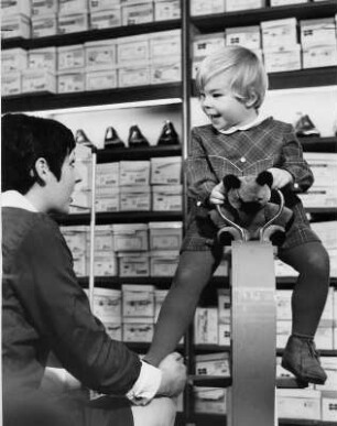 Schuhkauf. Einem Mädchen werden in einem Schuhgeschäft Schuhe anprobiert. Sie sitzt dabei auf einem speziellen Kindersitz