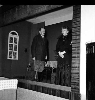 Laienspieler: Aufführung eines Theaterstücks: Bühne: Szenenbild: Frau und Mann stehen an Bühnenrand, dahinter Tisch