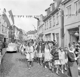 Kindervogelschießen: Umzug: Hindenburgstraße: kleine Mädchen mit Kränzchen im Haar, ältere Schülerinnen, Mädchen mit Blumenbögen, Jungen mit Blumenstäben und Fähnchen: an den Straßenrändern Zuschauer: Volkswagen "VW-Käfer": über Straße Wimpelgirlanden, an Fassaden Fahnenschmuck: an linker Straßenseite Friseursalon Lübcke, Gaststätte "Bierquelle": im Hintergrund Hagenstraße, Gaststätte "Stadt-Krug", Toreinfahrt Holz- und Baustoffhandlung Hugo Eggers, 30. Juni 1964