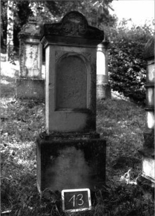 Grabstein 13 (Index: 100100940) Material: Sandstein Zustand: Schrifttafel fehlt Maße HxBxT: 173x43x19 cm Beschreibung: Stele, Sockel, verbreiterter Aufsatz mit eingezogenem Rundbogen, eingetieftes Schriftfeld mit Rundbogen Symbolik: Blattwerk im Aufsatz: Grabstein 13