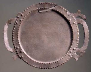 Runder Teller aus flachem Blech mit zwei Henkeln in Schleifenform; der Rand mit kleinen Buckeln verziert