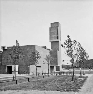 Klosterbergen: Täby-Platz: vorn eingezäunte Grünanlage mit Bäumen: links dahinter evangelische Nathan-Söderblom-Kirche: rechts dahinter drei Personengruppen auf Weg: hinten rechts Reihenhäuser