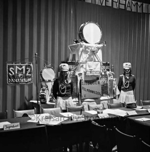 Oldesloer Spielmannszug e.V.: Jahresversammlung: Hotel Oldesloer Hof: leerer Vorstandstisch mit Platzkärtchen der Funktionsträger: hinten Ausstellung von Instrumenten und Standarten: daneben zwei Mädchen in Uniformen des Spielmannszugs: Aufnahme unscharf, 15. Februar 1975