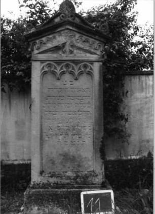 Grabstein 11 (Index: 99936899) Material: Sandstein Zustand: verwittert und beschädigt mit Textverlust Maße HxBxT: 168x56x28 cm Beschreibung: Stele, profilierter Sockel, giebelartiger Abschluß mit Verdachung mit Volutenaufsatz mit Mittelakroter, Schriftfeld eingetieft Symbolik: Maßwerk, gestelztes Rundbogenfries: Grabstein 11