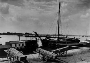 Hafen bei Emmerich am Niederrhein, der letzten großen deutschen Stadt vor der deutsch-niederländischen Grenze. Lastkähne auf dem Rhein. Ein Segelschiff und Lastkähen liegen am Kai. Einrädige Karren mit Ziegeln stehen am Ufer