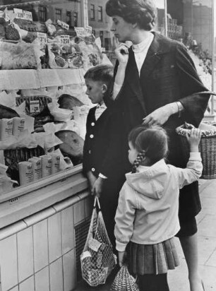 Eine Frau mit ihren Kindern beim Einkauf. Sie prüft die Preise in den Auslagen kritisch. Aufgenommen 1961