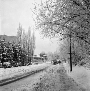 Hansdorfer Landstraße: rechts und links Bäume: hinten links Wohnblock und Einfamilienhäuser: hinten Mitte parkende Pkws, daneben zwei Personen: Winter