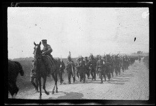 Nicht identifizierbare Ortschaft in Nordfrankreich oder Belgien: Soldaten mit Pickelhaube und geschultertem Gewehr auf einer unbefestigten Landstraße, im Vordergrund ein Offizier zu Pferde, dahinter ein Fahnenträger