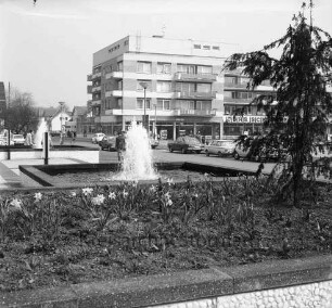 Rathausvorplatz: Blumenbeet mit Narzissen und Nadelbaum, Springbrunnen, Lampen: Reeshoop [später Manfred-Samusch-Straße], Autos, Straßenlampen: hinten Wohn- und Geschäftsblock der nördlichen Randbebauung von Rathausplatz, mit Geschäften Schuhhaus "Bata", "Johs. Schmidt Markt"