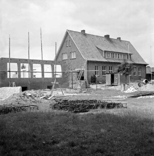 Volksschule: Rönnbaum: Erweiterungsbau: links errichtete Grundmauern mit Gerüst: rechts Schulgebäude: davor Baumaterial und Baumaschinen
