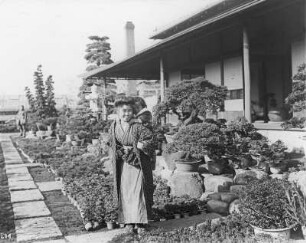 Yokohama. Botanischer Garten. Japanerin mit Kind auf dem Rücken im Bonsaigarten vor einem Gebäude