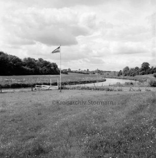 Trave: Bootsanleger unterhalb des Ortes: vorn Wiese mit Fahnenmast: im Hintergrund Knicklandschaft