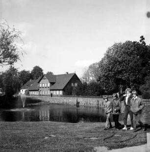 Dorfstraße: Dorfteich: vorn rechts Gruppe Jugendlicher: dahinter Personengruppe: links Birke: dahinter Springbrunnen: hinten Bäume und Häuser