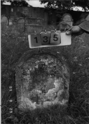 Grabstein 135 (Index: 100107384) Material: heller Sandstein Zustand: stark verwittert und bewachsen, völliger Textverlust Maße HxBxT: 58x46x14 cm Beschreibung: Stele mit Rundbogenabschluß, Schriftfeld eingetieft: Grabstein 135