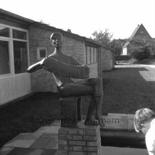 Ahrensburg: Reeshoop: vor Fertigstellung: Außenaufnahme: Kunst am Bau, Bronzeplastik "Ziehharmonikaspieler" von Bildhauer Karlheinz Goedtke, an Wasserbecken: Kopf von Junge: Rasen: hinten Wohnhaus