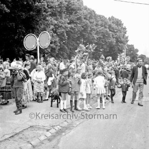 Kindervogelschießen: Festumzug: Klassen mit Blumenstöcken: Schulkinder in Kostümen: rechts Zuschauer: links Verkehrsschild: hinten Bäume