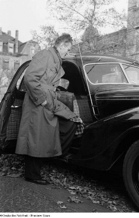 Ein Mann an einem PKW präsentiert einen Trenchcoat des VEB Bekleidungswerk Leipzig