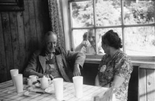 Kaffeepause : Zwei Personen an einem Kaffeetisch vermutlich in der Wurzhütte. Ein Mann schaut durch ein Fester in die Hütte