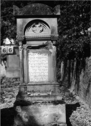 Grabstein 66 (Index: 100086522) Material: Sandstein mit weißer Marmorplatte Zustand: verwittert Maße HxBxT: 179x66x25 cm Beschreibung: ädikulaartige Stele mit dünnen Halbsäulen, getreppter Sockel, Segmentbogengiebel mit Eckakroteren, eingetieftes Schriftfeld rechteckig mit aufgesetztem Rundbogen Symbolik: Maßwerk im Rundbogen, drei achtzackige Sterne, Sternfries, eingemeißelte Verzierungen: Grabstein 66