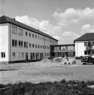 Ahrensburg: Hermann-Löns-Straße: Erweiterungsbau: vor Fertigstellung: Außenansicht: Herrichten des Vorplatzes: Arbeiter beim Verlegen von Betonplatten, Kleintransporter: hinten Bildmitte Verbindungstrakt, rechts Altbau