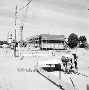 Langnese: Gewerbegebiet: Hammoorer Weg: Honigfabrikation: Betriebsgebäude in Bau: Rohbau mit Gerüsten, Baumaterialien: Baukran, Betonsilo: vorne Baugrube mit Fundament, Erdhaufen, davor Arbeiter bei Vermessung und Einrichten einer Baustelle, Planmappe auf Schubkarre, provisorische Baustromleitung: hinten weiteres Firmengabäude in Bau, Haufen Mauersteine: im Hintergrund Wohnhäuser