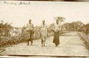 Drei Afrikaner, teils europäisch gekleidet, auf einem Weg an einem Fluss mit einem Geschütz im Hintergrund
