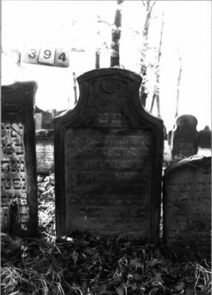 Grabstein 394 (Index: 100115694) Material: Sandstein Zustand: verwittert Maße HxBxT: 112x60x16 cm Beschreibung: Stele mit konkav eingezogenem Segmentbogenabschluß, eingetieftes Schriftfeld Symbolik: Blüte, Ranken: Grabstein 394