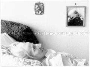 Kopf und Oberkörper einer im Bett liegenden alten Frau, dahinter an der Wand ein Babyfoto (Altersgruppe 18-21)