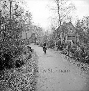 Lottbek: An der Lottbek: Brücke über die Lottbek an der Grenze zu Hamburg: rechts und links Büsche: rechts Ortseingangsschild für Hoisbüttel: in der Mitte Fahrradfahrerin: hinten rechts zwischen Büschen und Bäumen Haus
