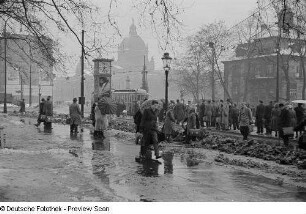 Menschen mit Regenschirmen warten an der Straßenbahnhaltestelle auf dem Martin-Luther-Ring, im Hintergrund das Reichsgericht